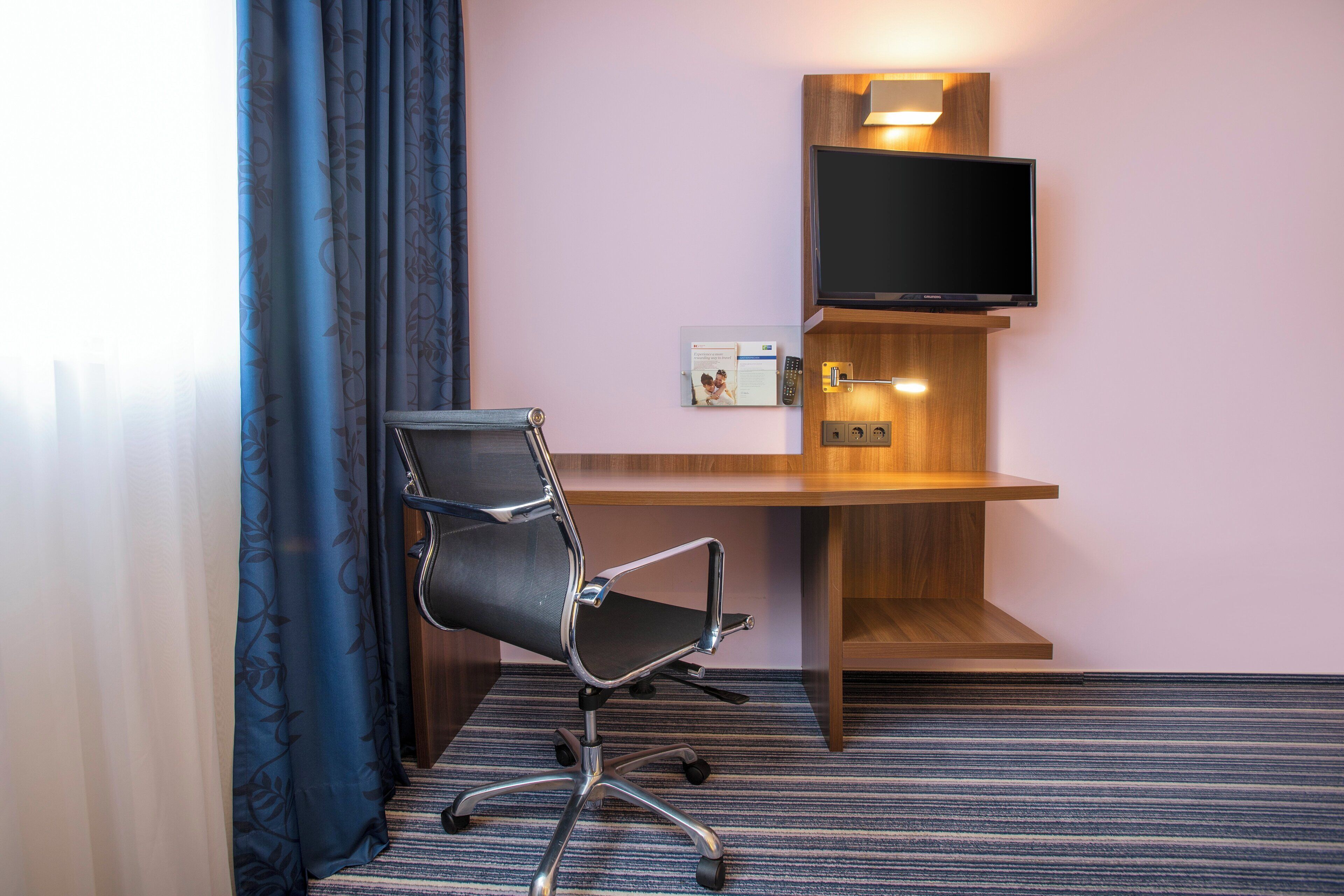 A hotel room with a desk, chair, television, and a power outlet.