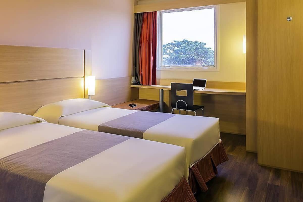A hotel room with two beds, a desk with a laptop, a chair, and a window with curtains.