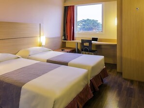 Ein Hotelzimmer mit zwei Betten, einem Schreibtisch mit einem Laptop, einem Stuhl und einem Fenster mit Vorhängen.