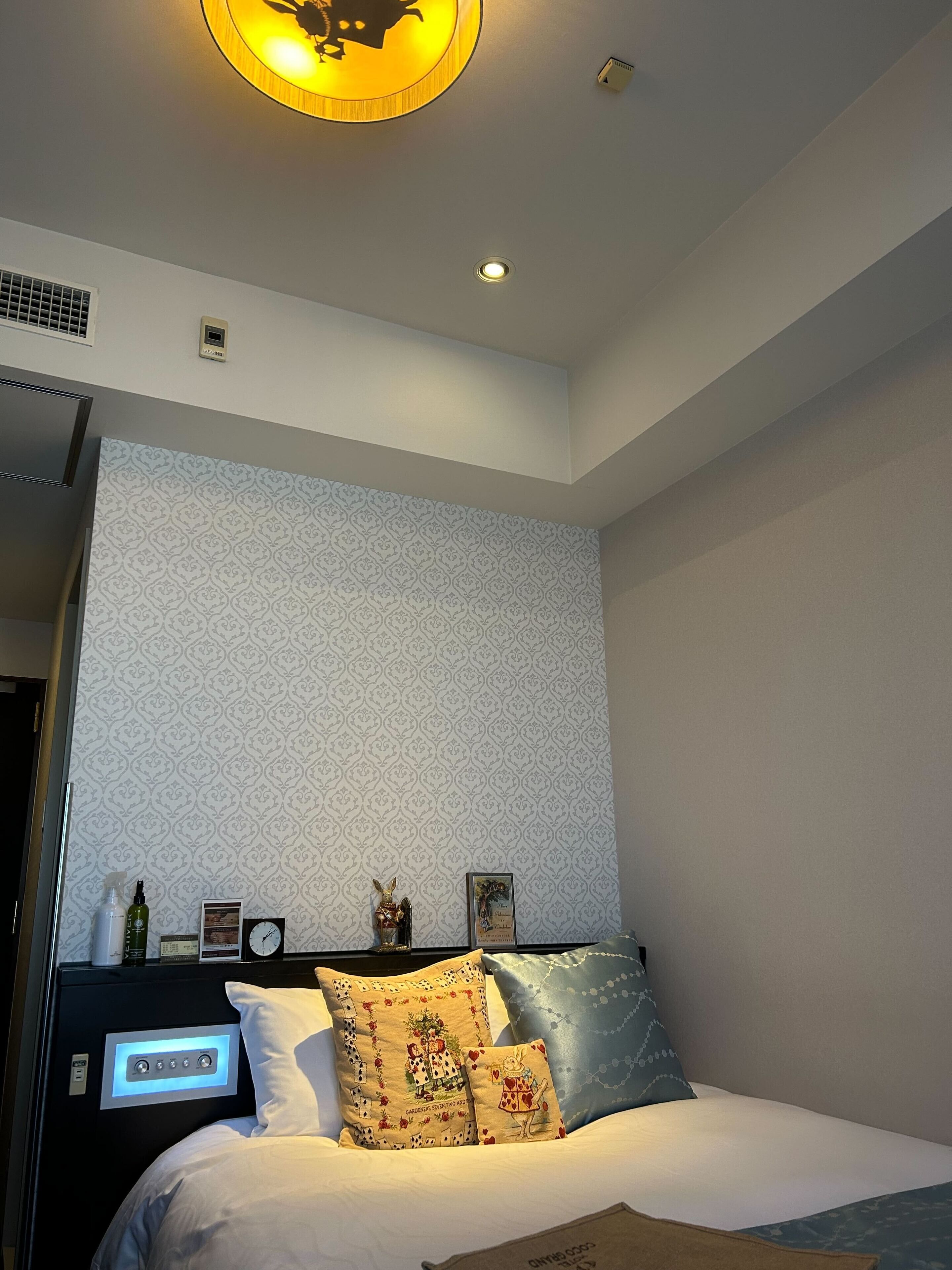 A modern bedroom with a patterned wallpaper, a bed with decorative pillows, a bedside table with a lamp, and a ceiling light fixture.