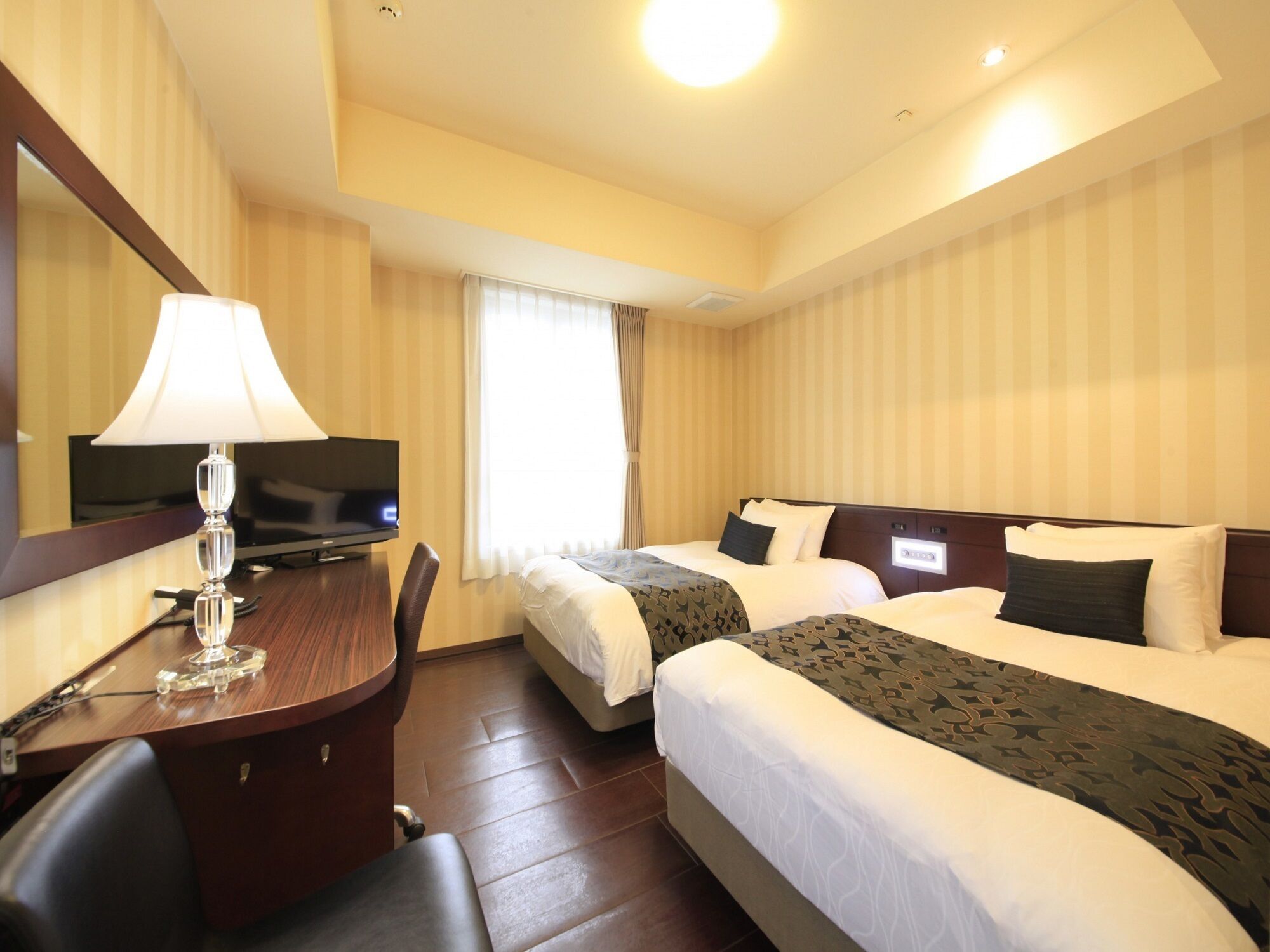 A hotel room with two beds, a desk with a lamp, a television, and a window with curtains.