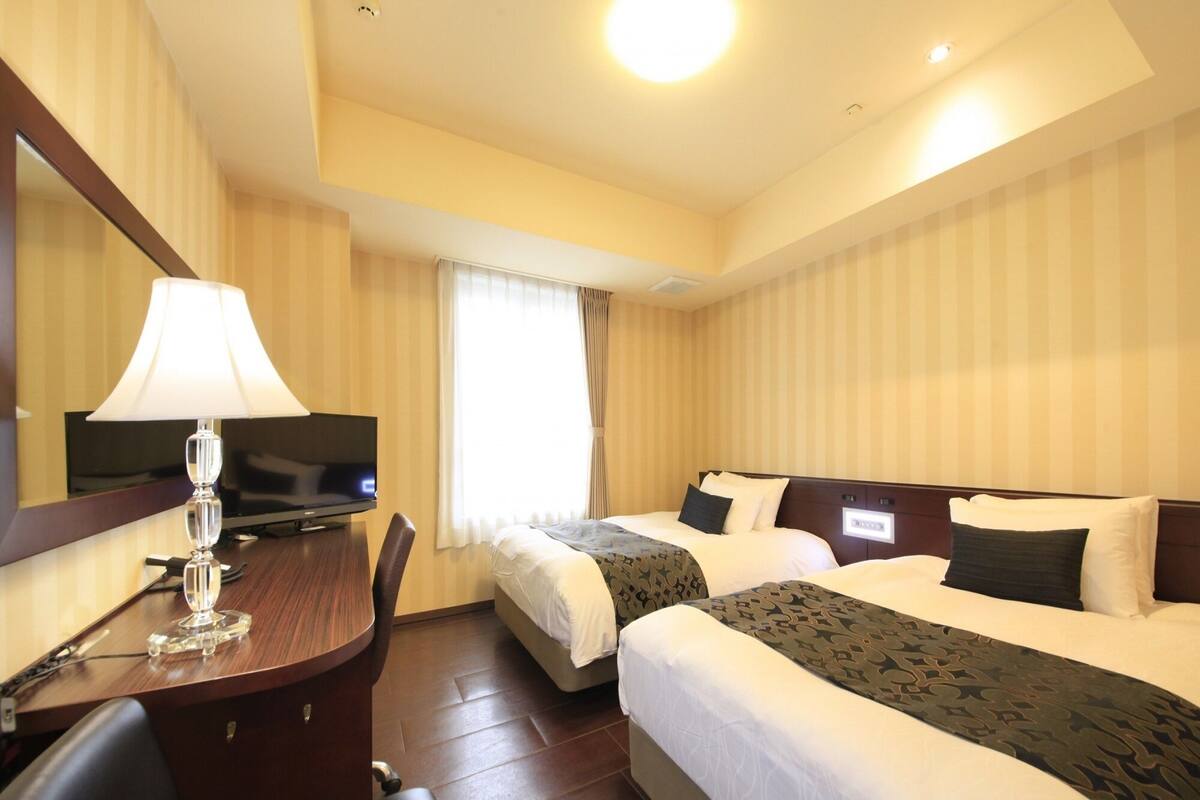 A hotel room with two beds, a desk with a lamp, a television, and a window with curtains.