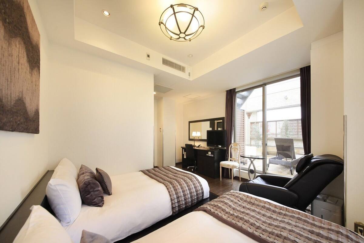 A hotel room with two beds, a desk, a chair, and a large window.