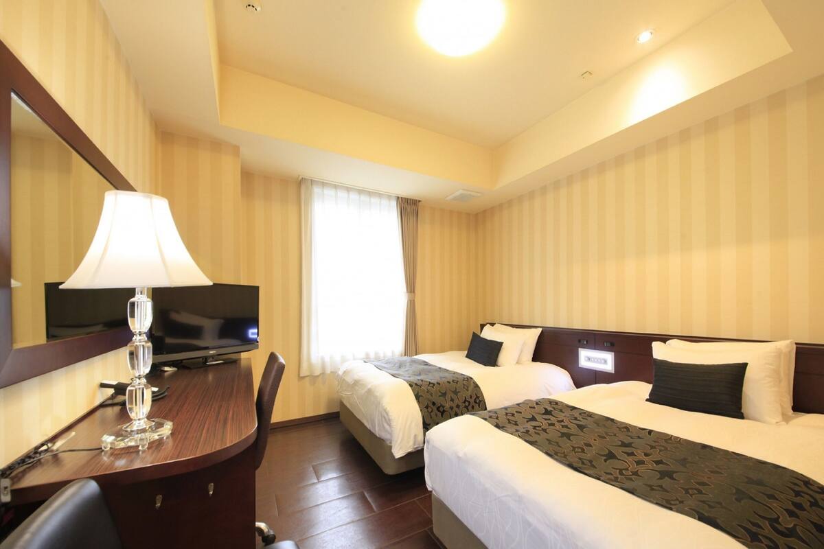 A hotel room with two beds, a desk with a lamp, a TV, and a mirror.
