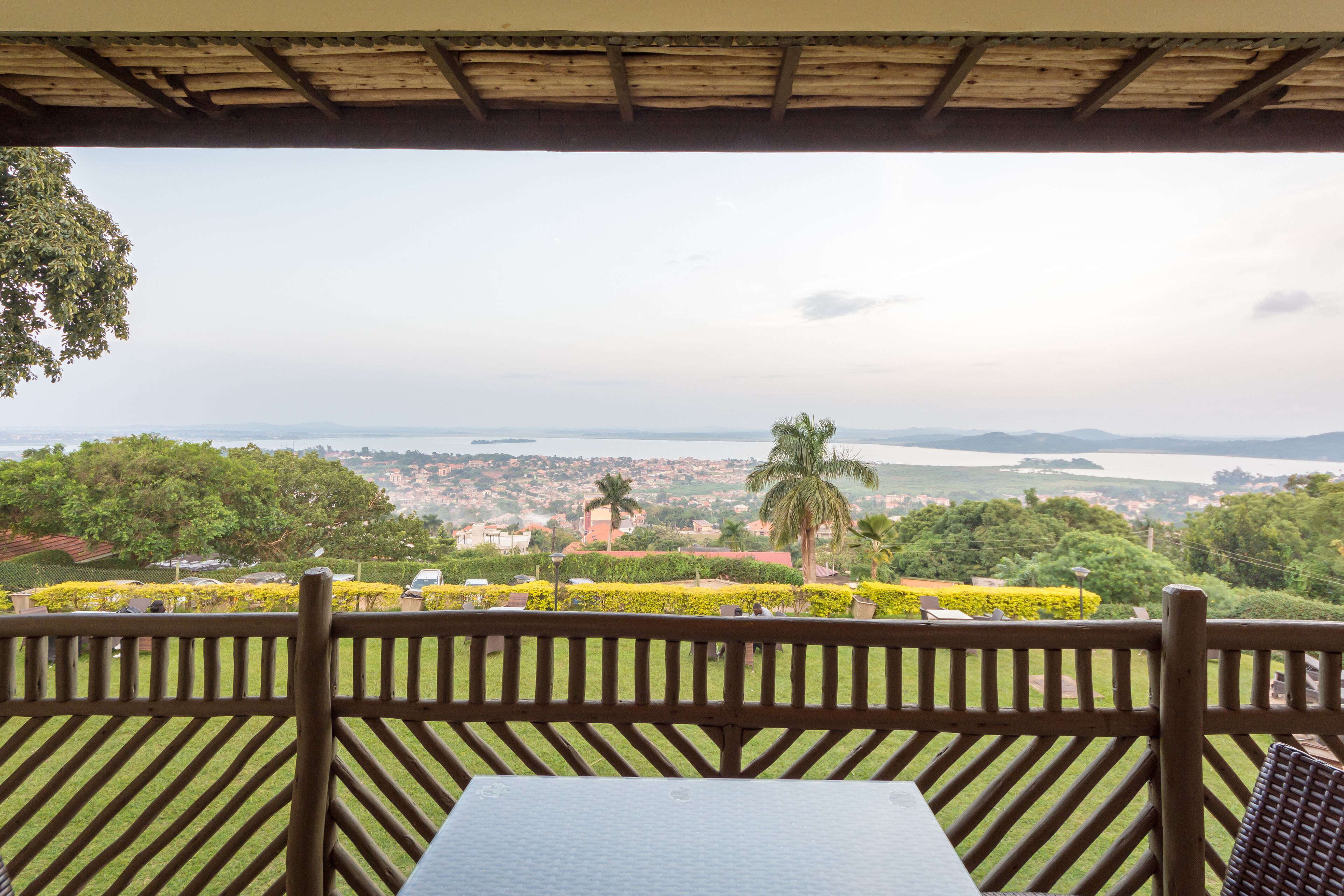 Une terrasse avec une table et des chaises, offrant une vue panoramique sur un paysage verdoyant, avec des palmiers et, au loin, sur une ville et une étendue d’eau.