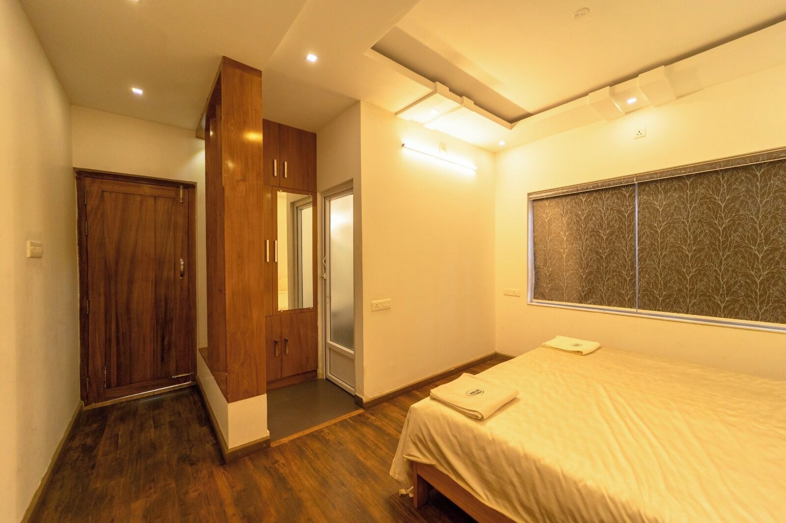 A bedroom with a wooden wardrobe, a bed with a white bedspread, and a window with a tree pattern.