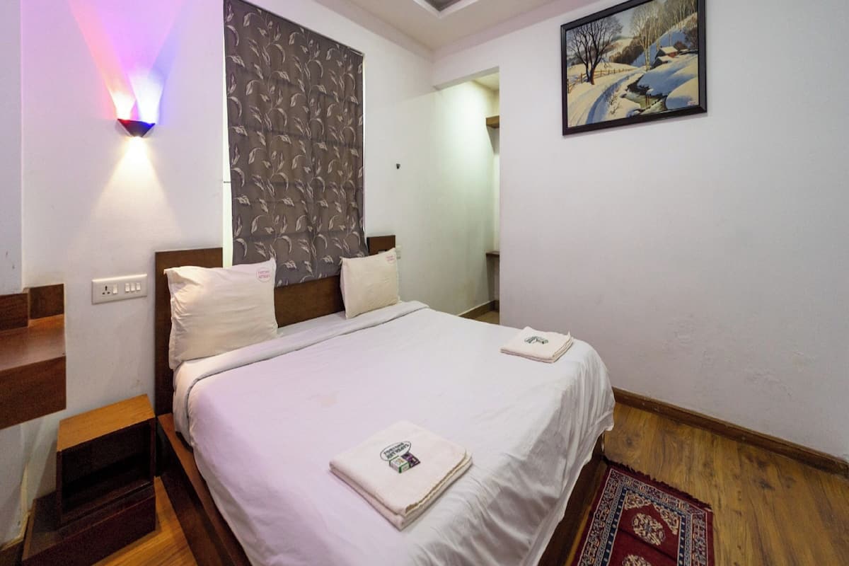 A hotel room with a bed, a nightstand, a wall-mounted light, a painting, and a red carpet.