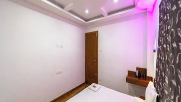 A bedroom with a bed, a wooden door, a small shelf, and a wall with a power outlet.