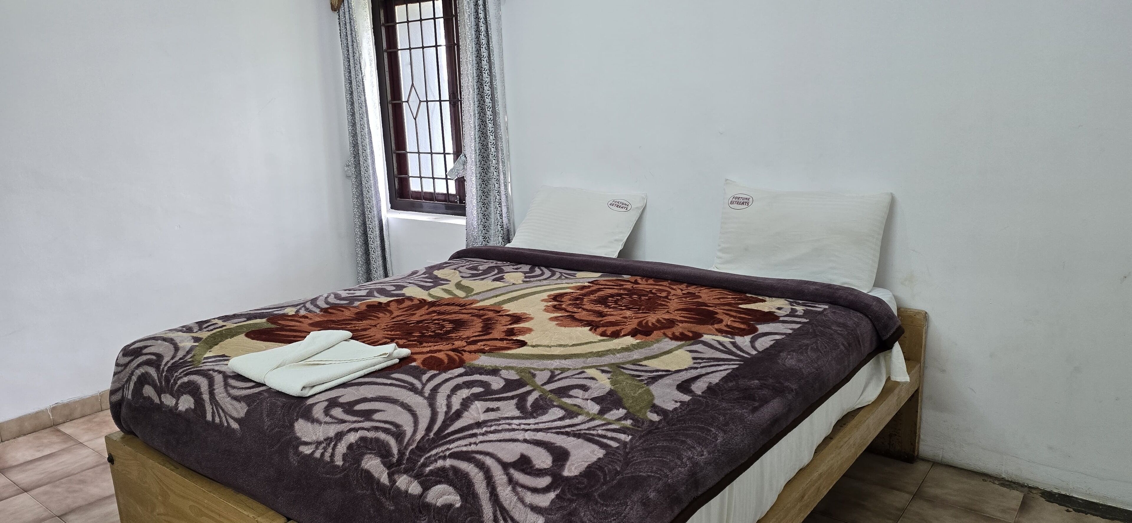 A neatly made bed with a floral bedspread, a white pillow, and a folded white towel on the side.