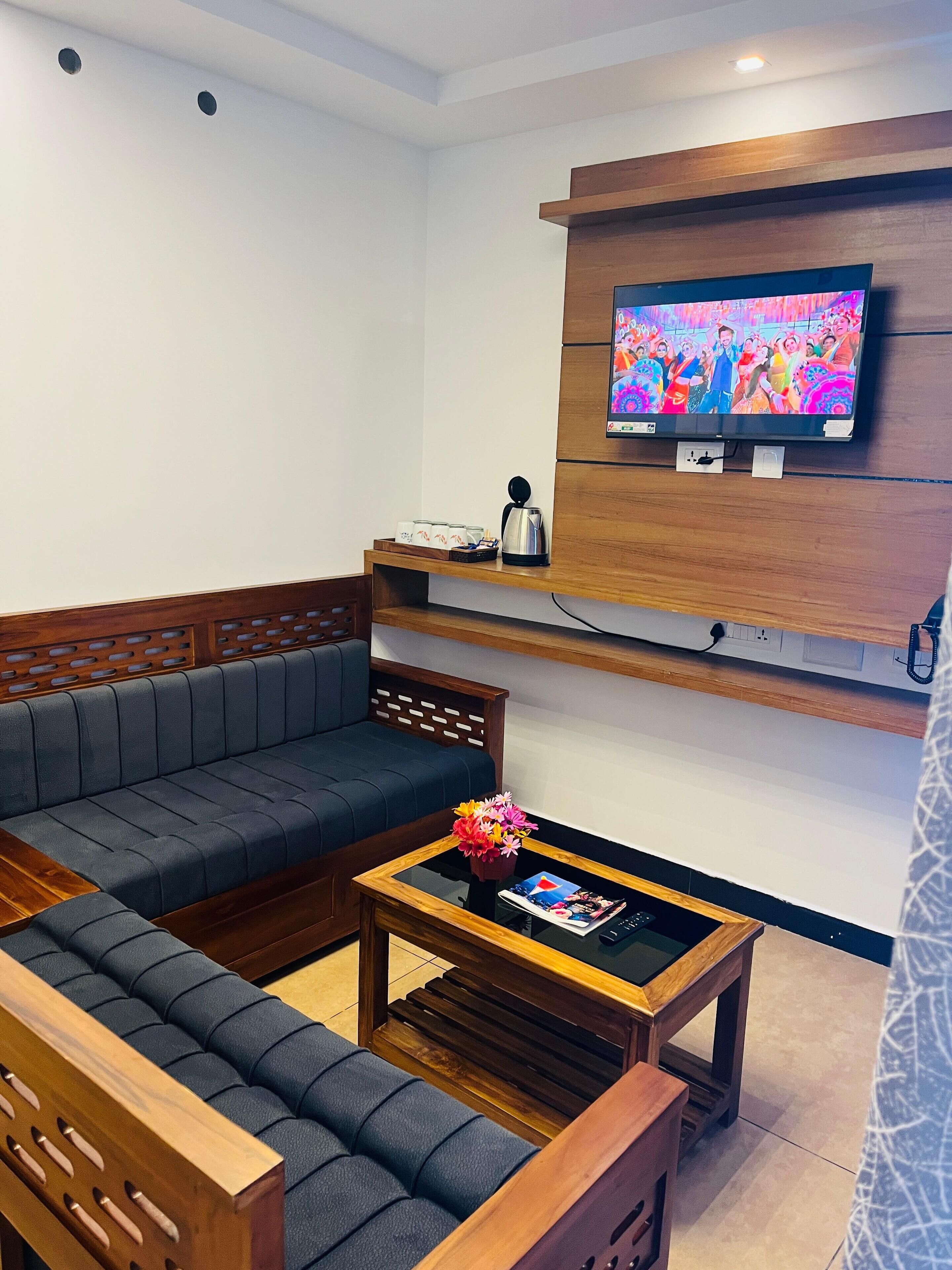 A modern living room with a sectional sofa, a coffee table with a magazine, a TV mounted on the wall, and a small shelf with a kettle and cups.