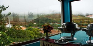 A view from a window overlooking terraced fields and a house in the distance, with a table set for tea and a vase of flowers on the windowsill. - Fortune Retreats (Ootacamund)