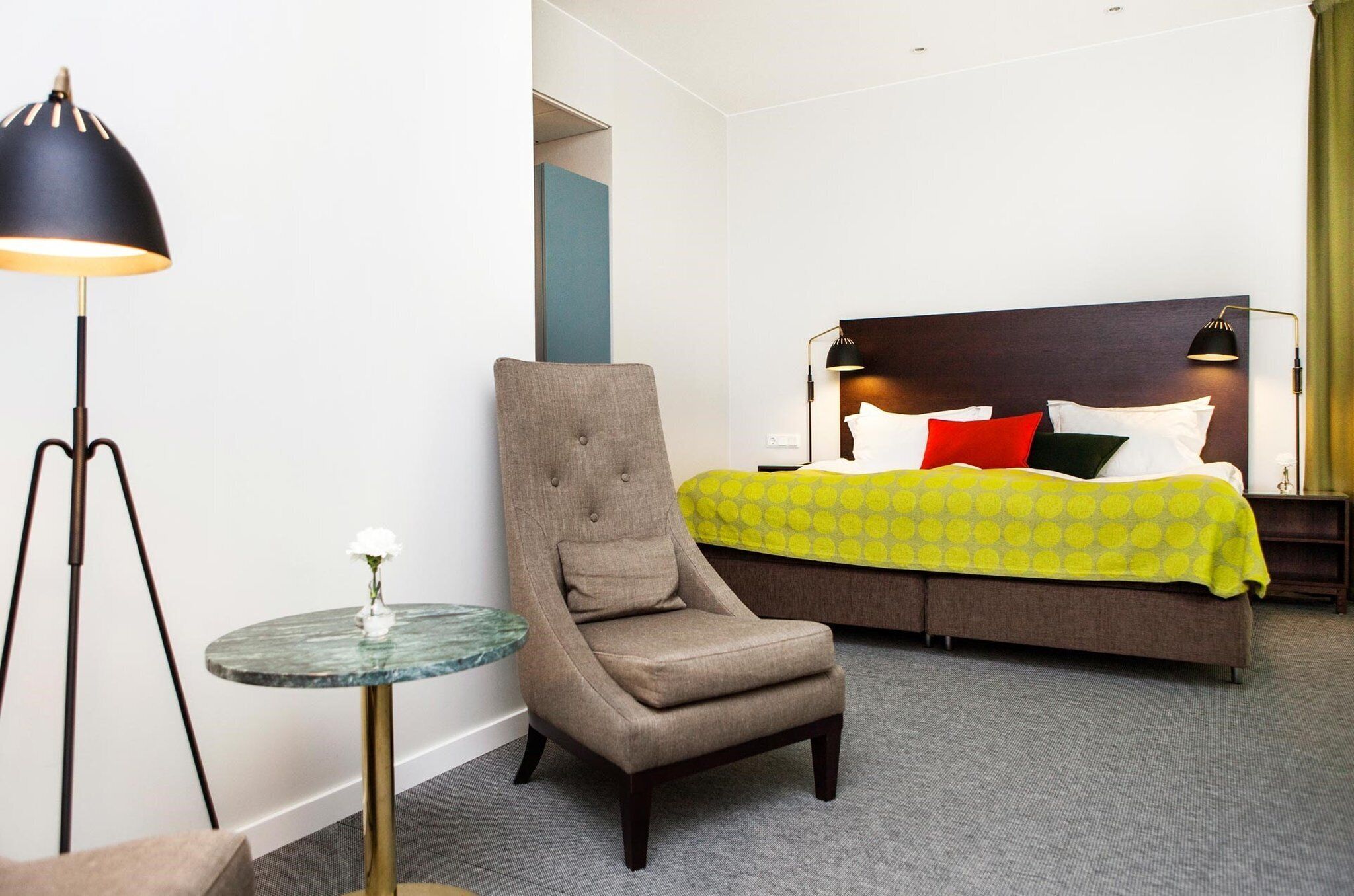 A hotel room with a bed, a chair, a small table, and a lamp.