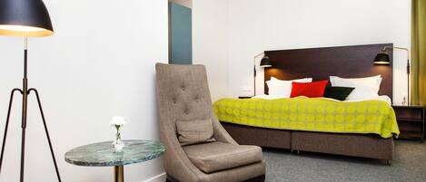 A hotel room with a bed, a chair, a small table, and a lamp.