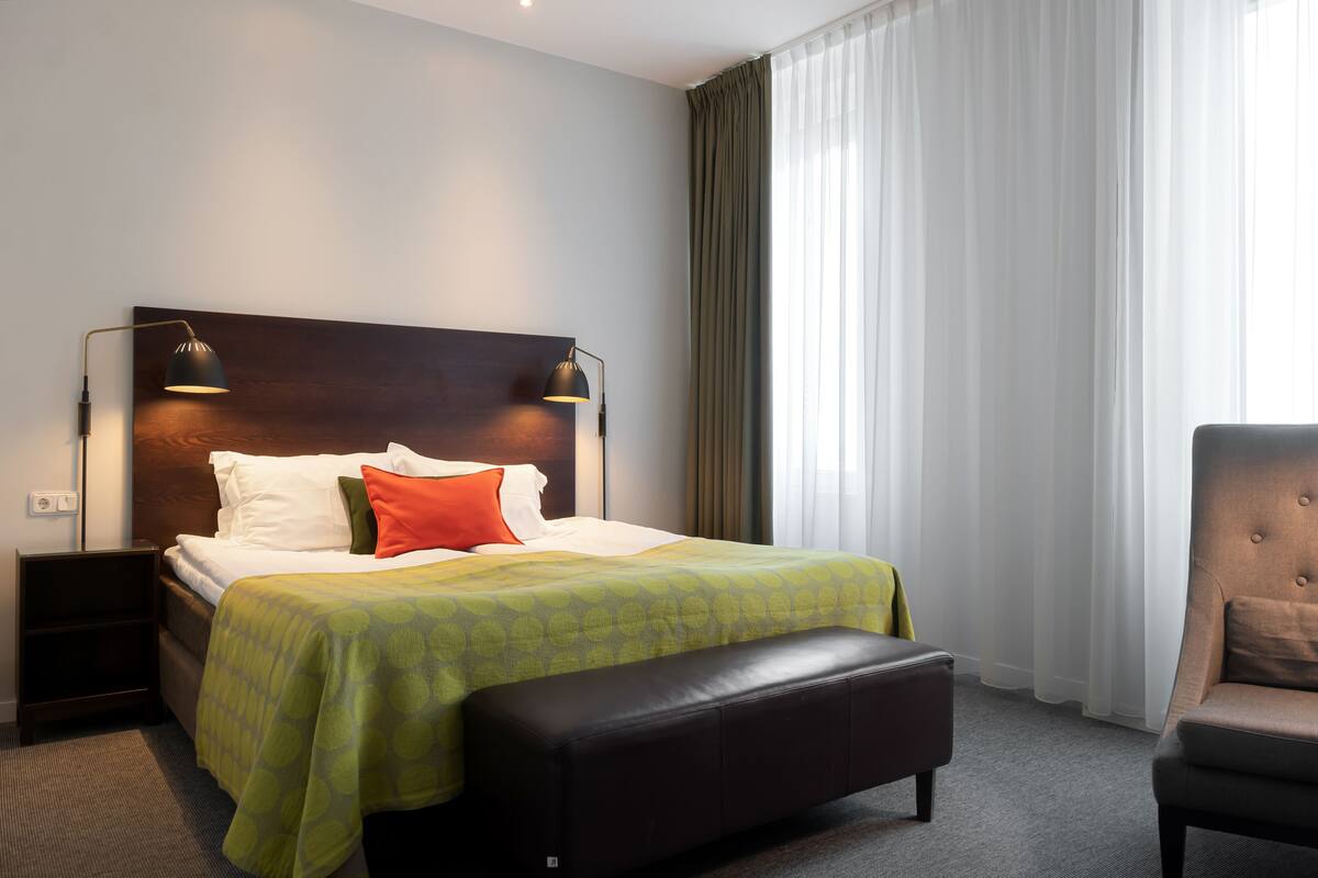 Ett hotellrum med en stor säng, två sänglampor, en bänk, en stol och utsikt över en byggnad genom tunna gardiner.