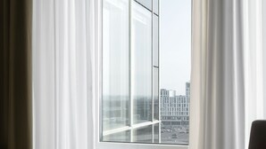 Una habitación con un ventanal grande, cortinas traslúcidas y vistas a un paisaje urbano, además de una silla marrón.