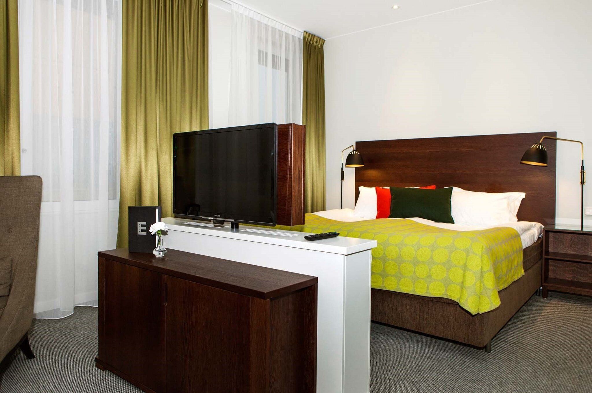 A hotel room with a bed, a TV on a desk, a lamp, and a remote control.