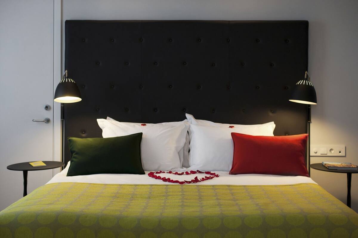 Ett hotellrum med en stoppad sänggavel, en säng med vita och röda kuddar, en grön pläd och två sänglampor.