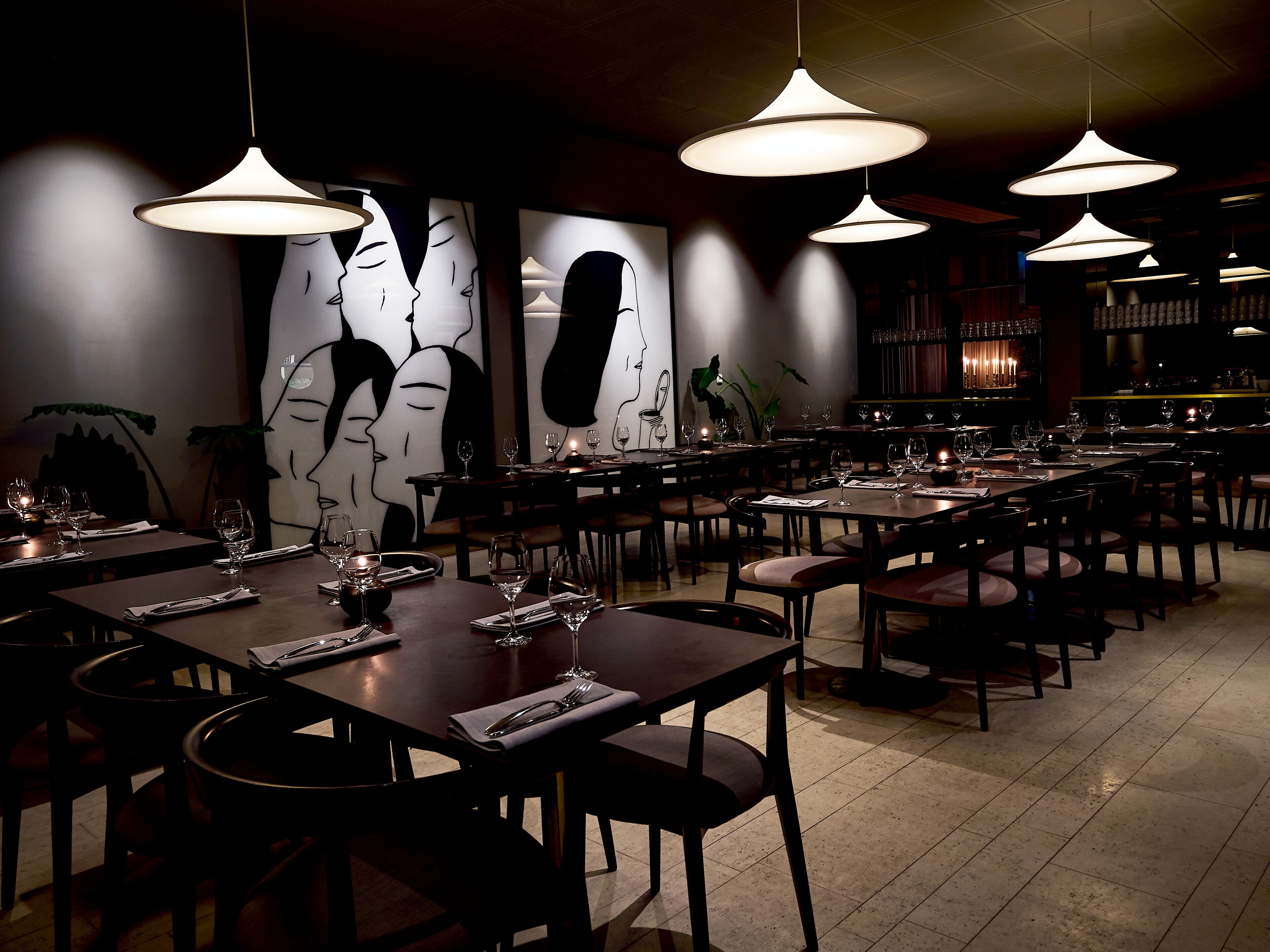 Een restaurant met meerdere tafels, elk gedekt met wijnglazen en bestek, en hangende lampen.