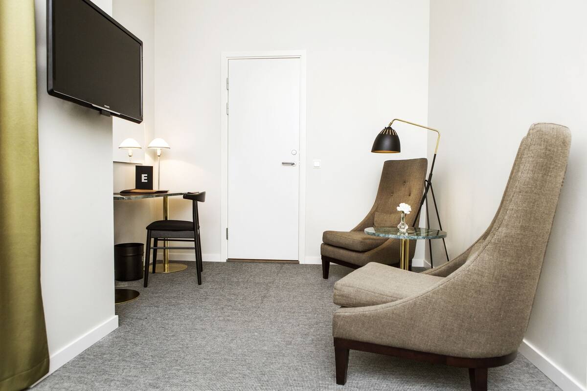 Ett modernt hotellrum med en platt-TV som är monterad på väggen, ett litet bord med en lampa, en bekväm fåtölj och en dörr.
