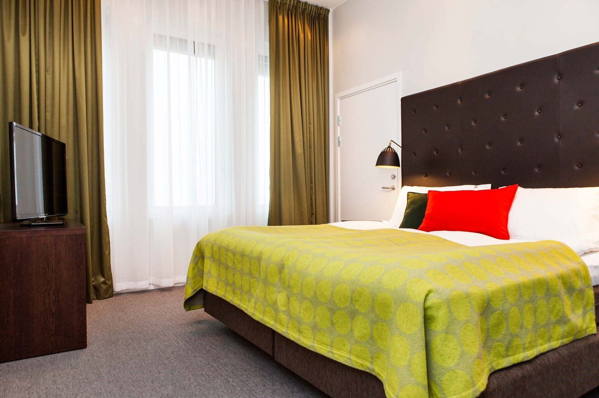 Una habitación de hotel con una cama, un televisor sobre un soporte de madera y una ventana con cortinas.