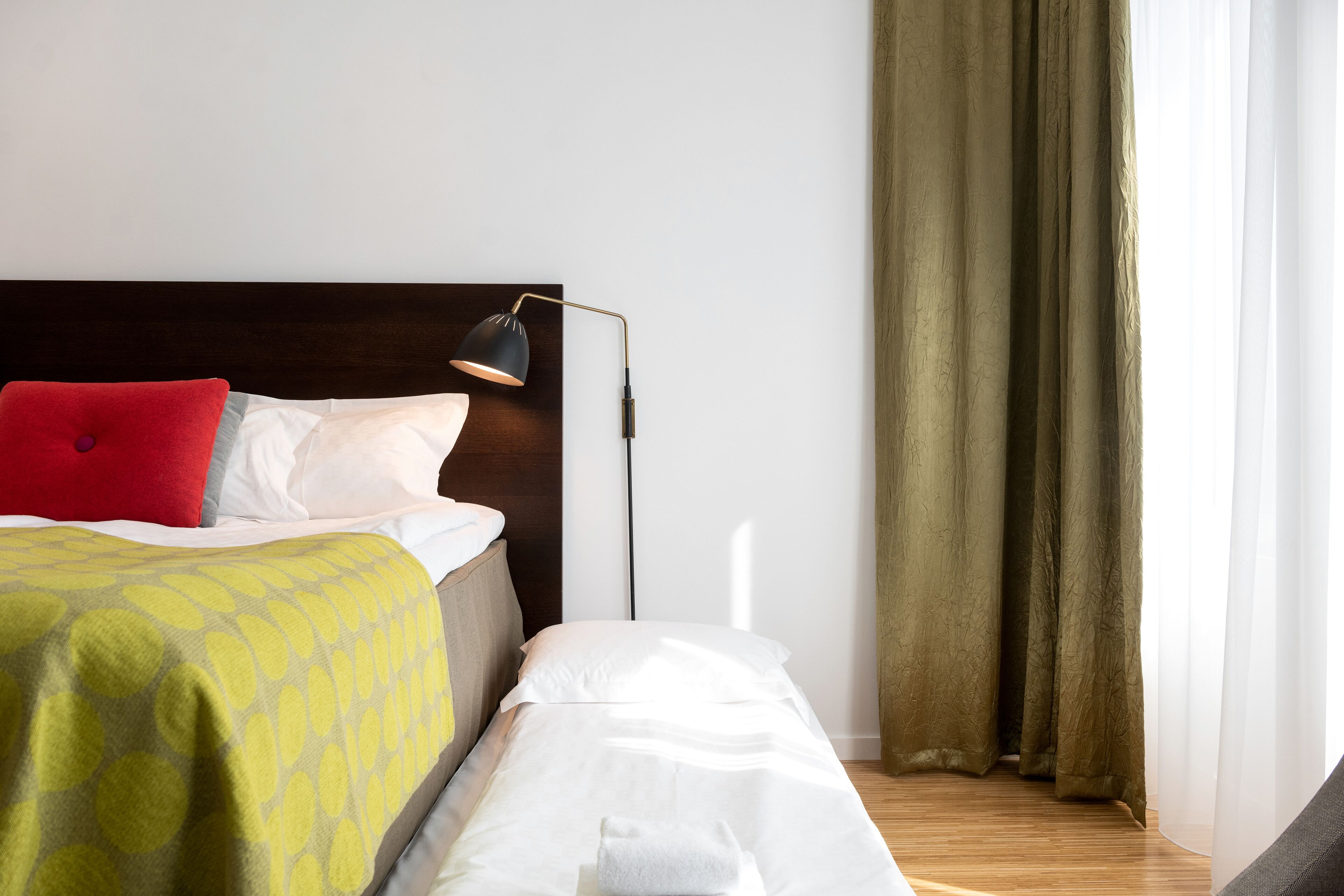 A neatly made bed with a red pillow, a yellow patterned blanket, and a black desk lamp.