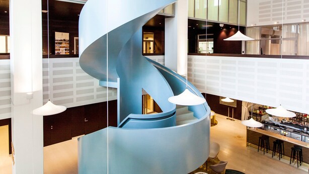 Un interior moderno con una escalera espiral azul, zonas de estar y un bar/lounge con techos altos y ventanas amplias.