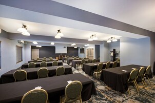 Un salón de conferencias con varias mesas y sillas, equipado con luces en el techo y una pantalla de proyector.