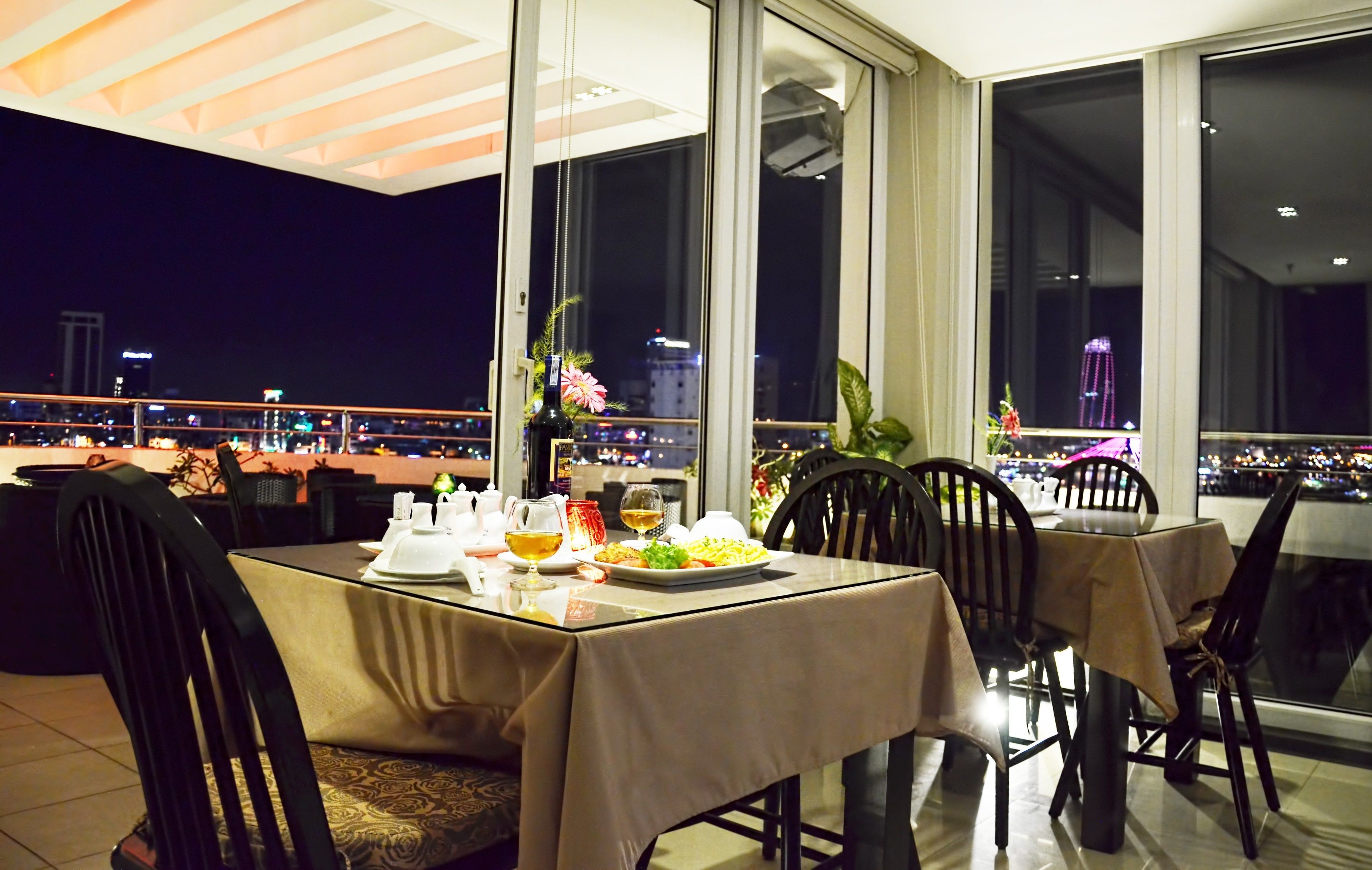 A dining area with a table set for two, a vase with flowers, and a city view through large windows.