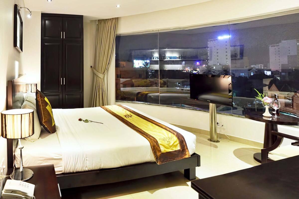 Une chambre d’hôtel avec un grand lit, un bureau, une chaise, une armoire et une grande fenêtre donnant sur la ville.