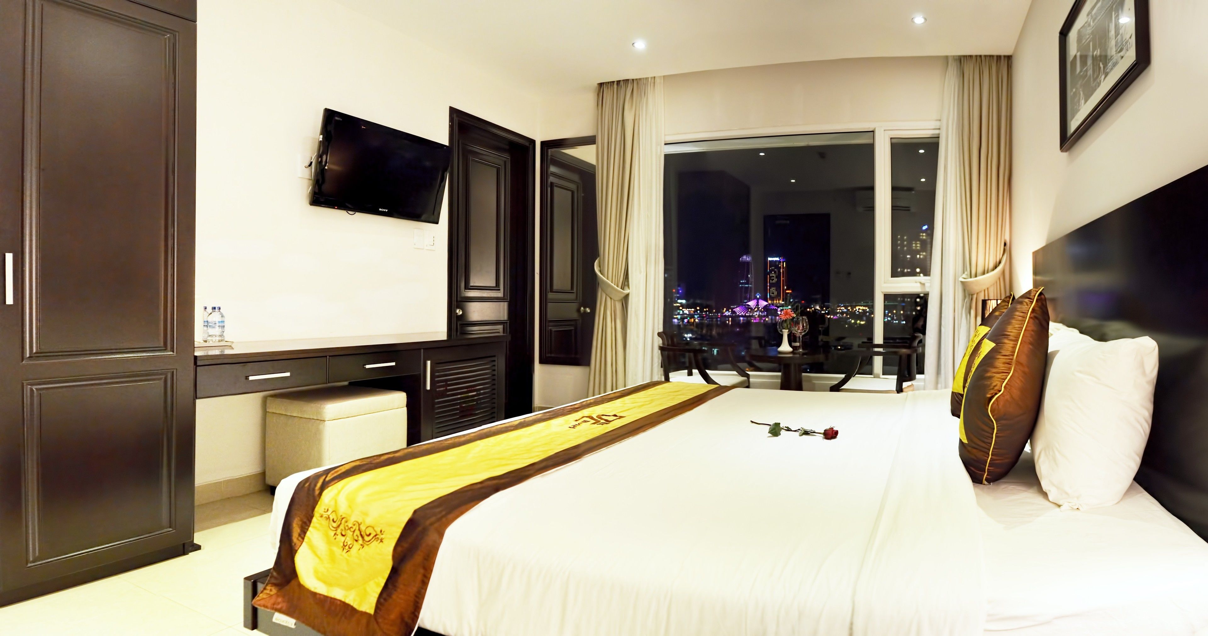 A hotel room with a bed, a TV, a desk, and a view of the city at night.