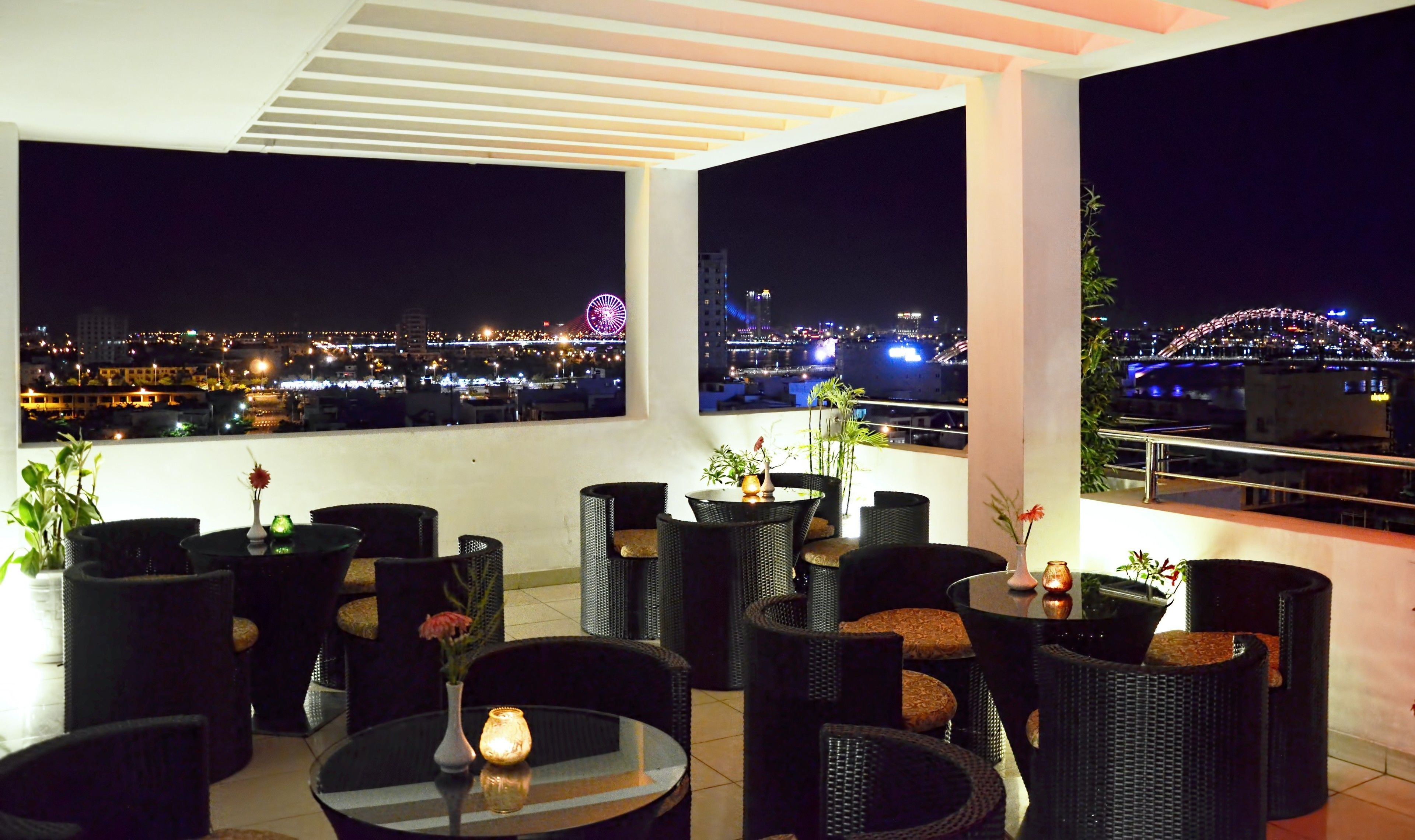 Una terraza en la azotea con mobiliario de mimbre, una pequeña mesa con una vela y vistas a un paisaje urbano nocturno.