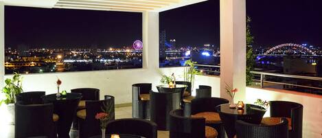 Una terraza en la azotea con mobiliario de mimbre, una pequeña mesa con una vela y vistas a un paisaje urbano nocturno.