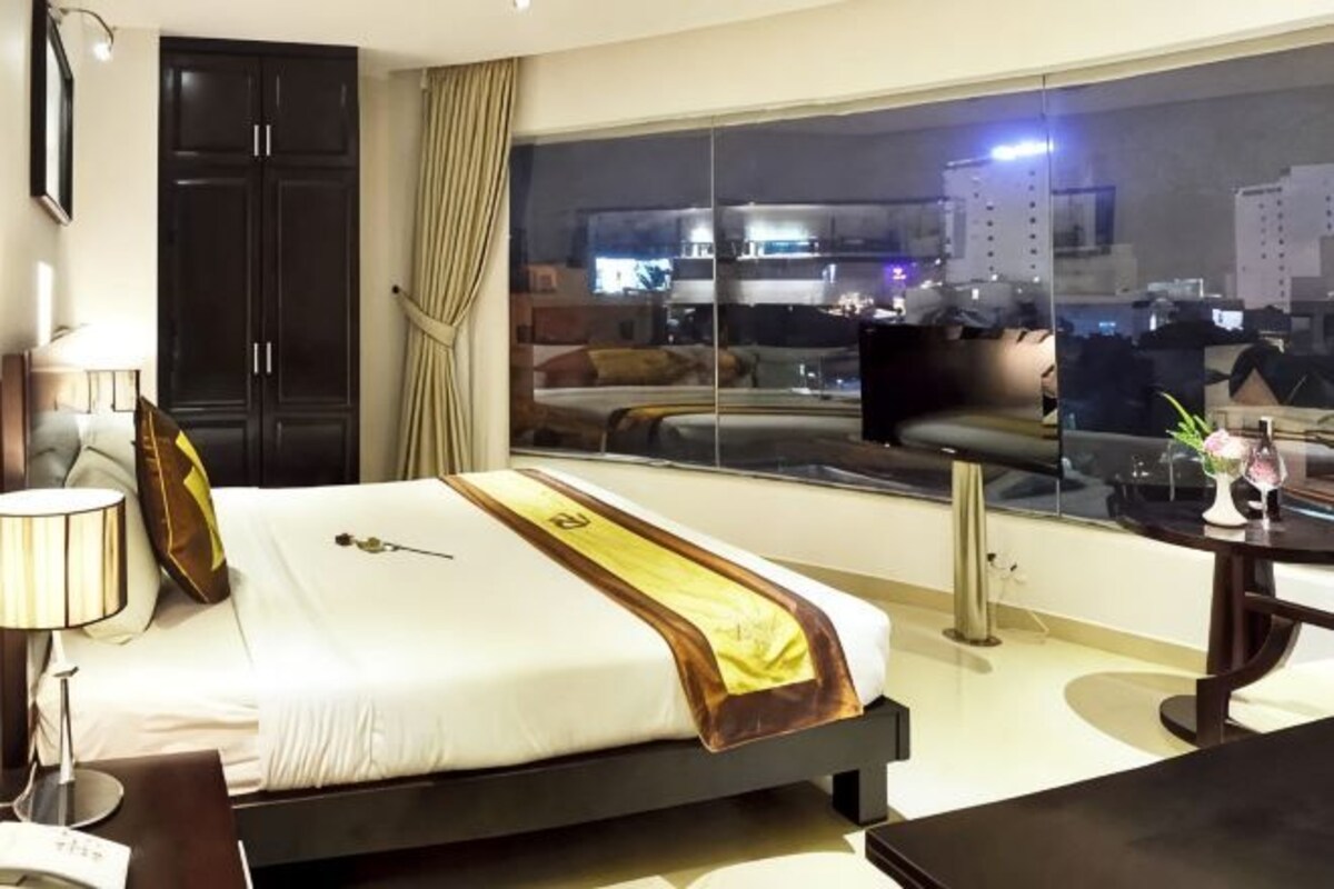 A hotel room with a bed, a desk, a chair, a window with a city view, and a large mirror.