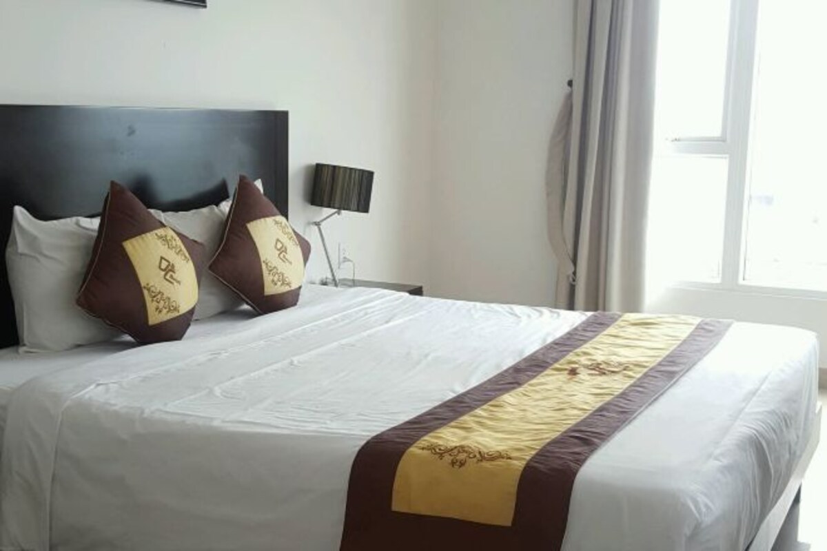 A neatly made bed with a white and brown bedspread, two decorative pillows, a bedside lamp, and a window with curtains.