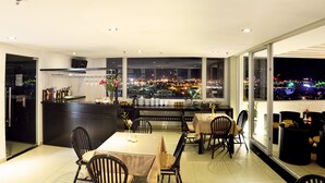 Un comedor con vistas a un paisaje urbano nocturno, que incluye una barra con botellas y vasos, y una mesa lista para dos personas.