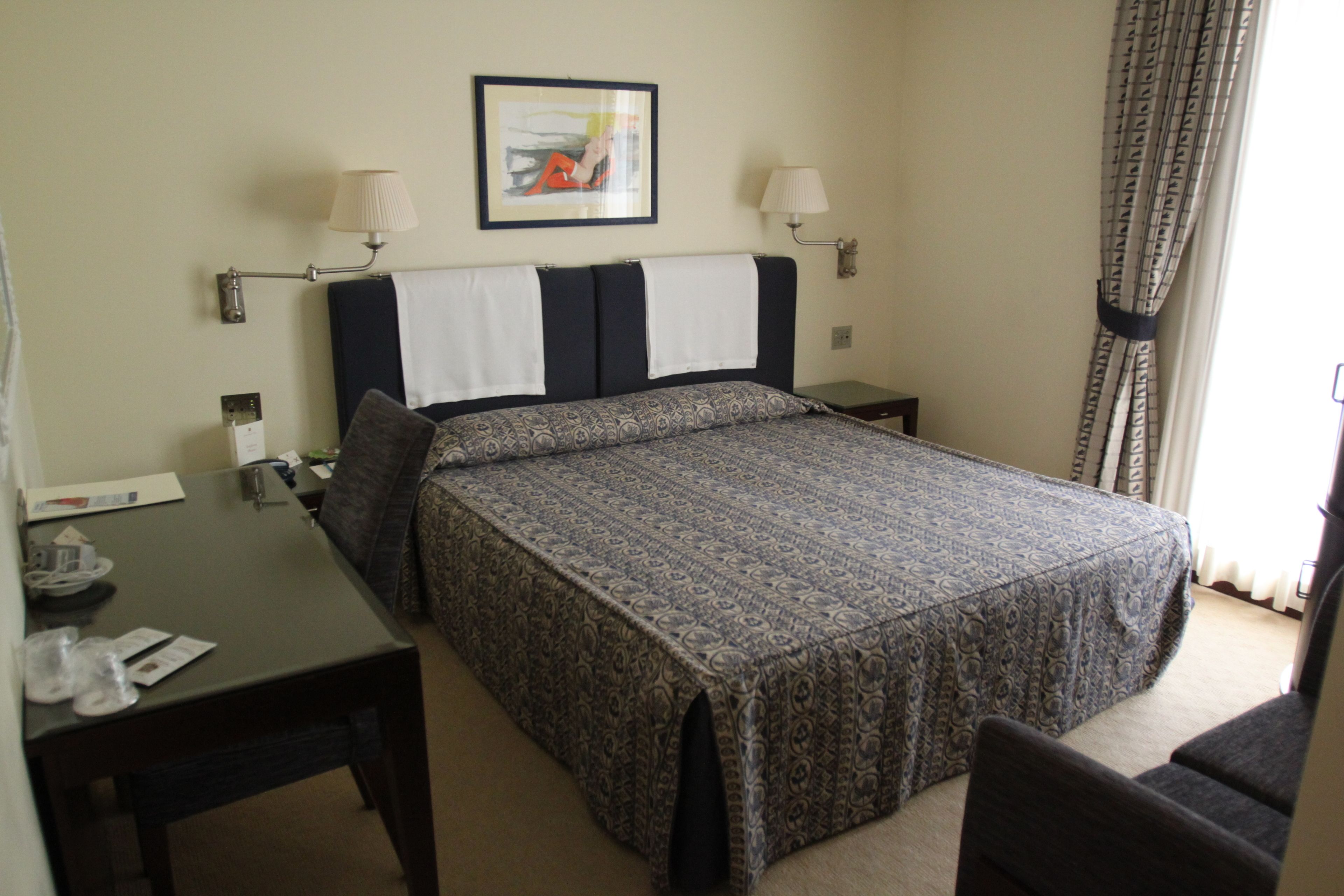 A hotel room with a bed, a desk with a chair, a lamp, and a framed picture on the wall.