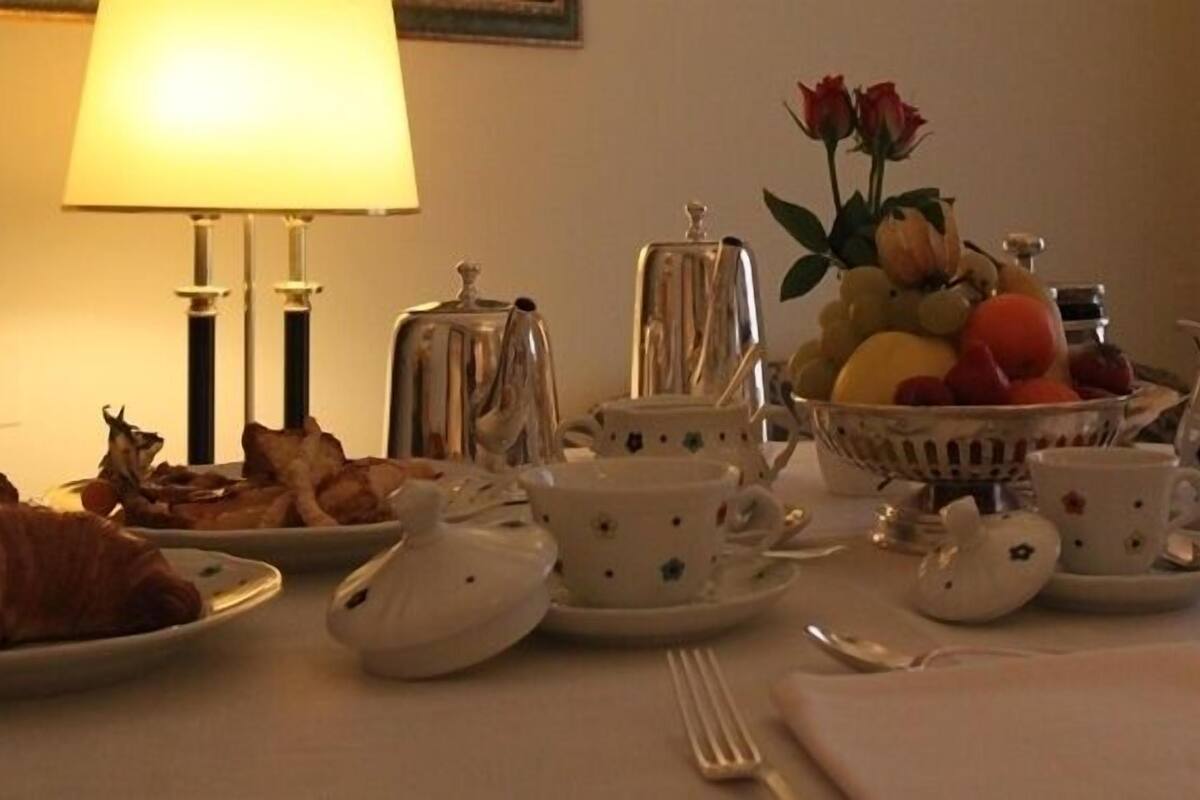 A breakfast setting with a table set for two, including plates of food, cups, a teapot, and a fruit bowl.