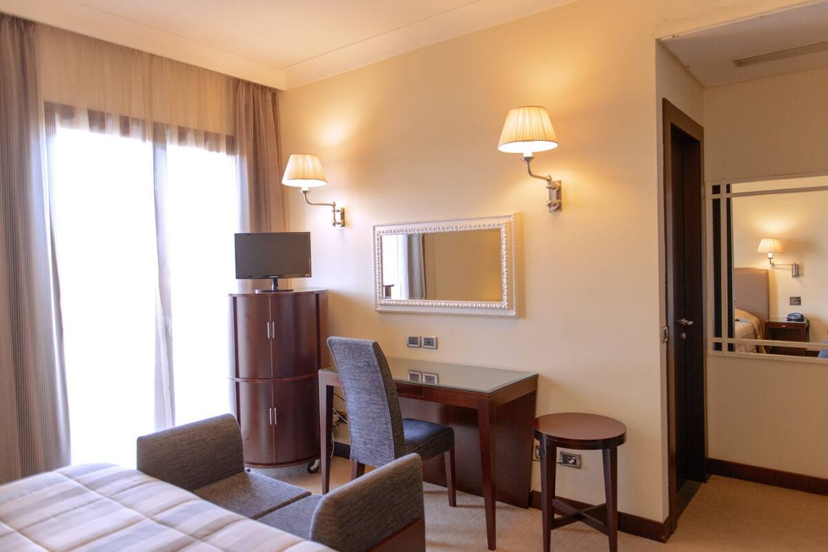 A hotel room with a bed, a desk, a chair, a TV, a mirror, and two wall-mounted lamps.