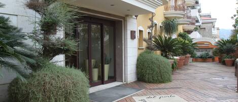 Entrada de hotel con un camino de ladrillo, plantas en macetas y felpudo con el nombre del hotel.
