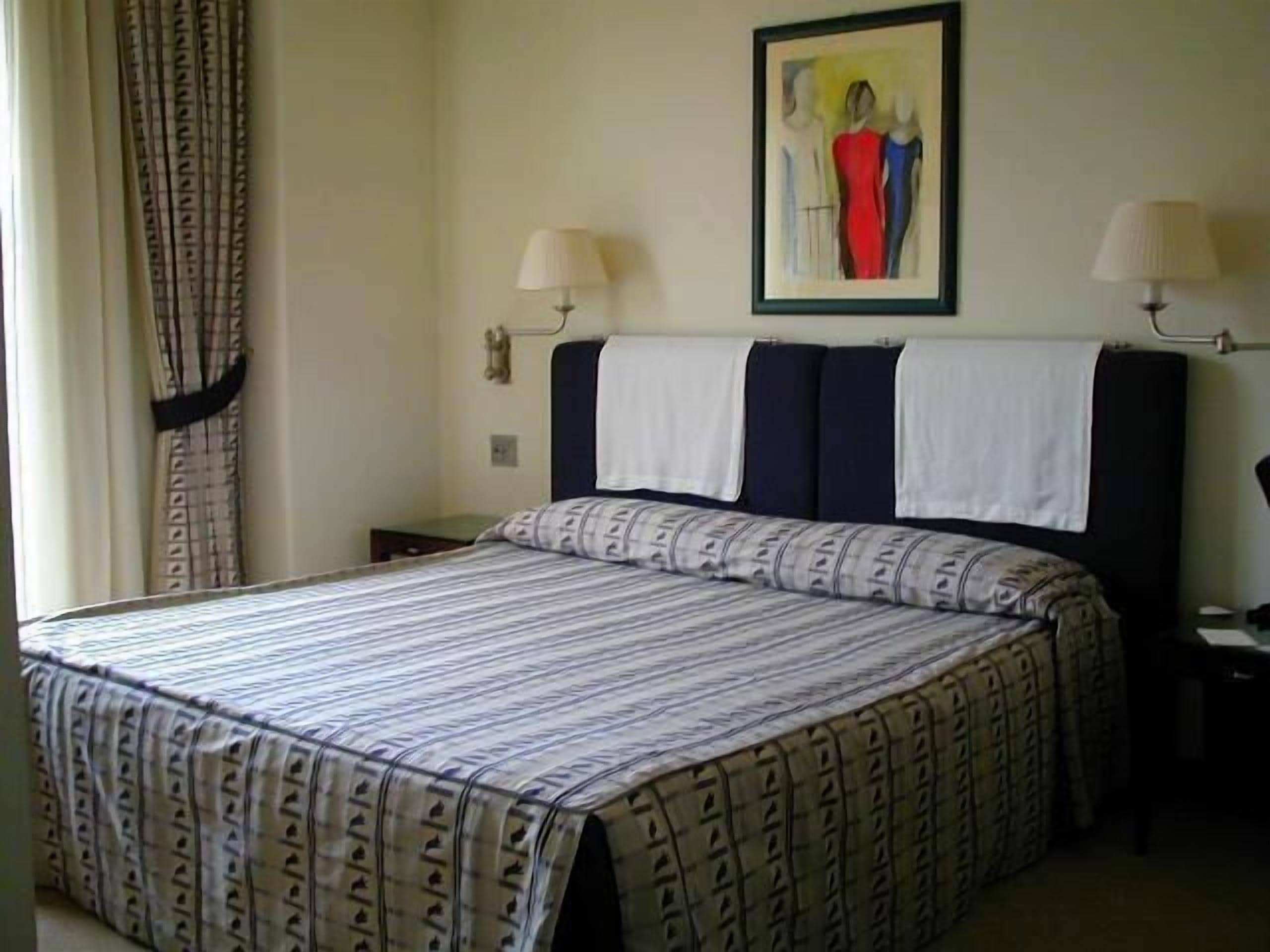 A hotel room with a bed, two bedside lamps, a painting on the wall, and a window with curtains.