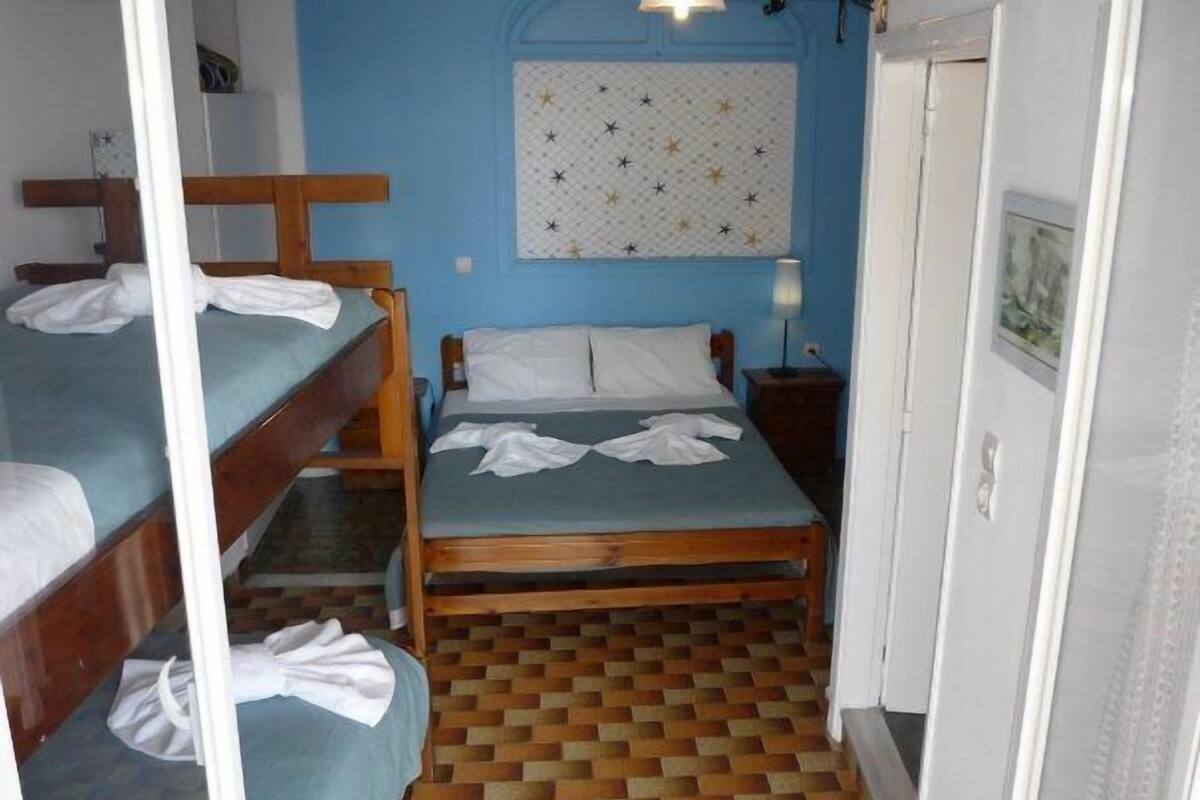 A dormitory room with two bunk beds, a single bed, a patterned wall hanging, a nightstand with a lamp, and a tiled floor.
