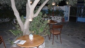 Ein Außenbereich mit Sitzgelegenheit, einem runden Tisch, Stühlen, einem Baum mit hängenden Laternen und einer Steinsäule im Hintergrund.