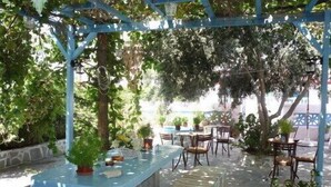 Ein Sitzbereich im Freien mit Tischen und Stühlen, umgeben von Grünpflanzen und einer blauen Pergola.