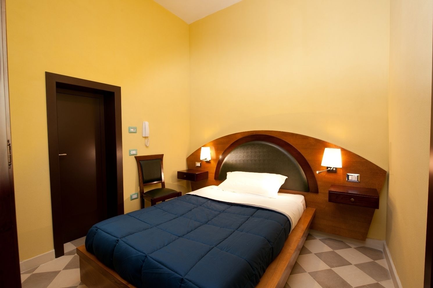Ein Hotelzimmer mit einem Bett, einem hölzernen Kopfteil, zwei Nachttischen mit Lampen sowie einer Kommode und einer Tür.