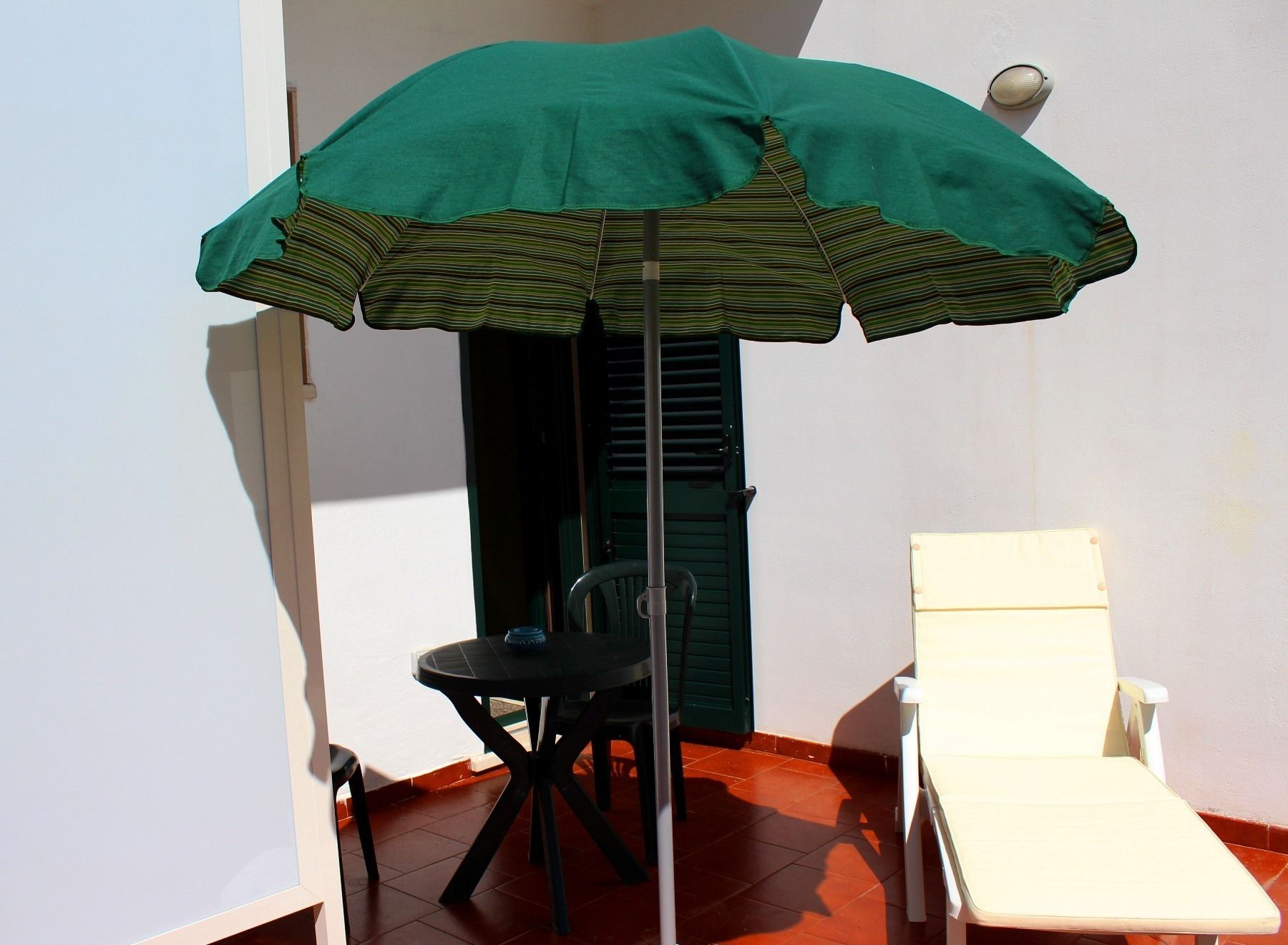 Un petit espace extérieur aménagé avec des sièges, comprenant un parasol à rayures vertes, une chaise noire et une chaise longue blanche.