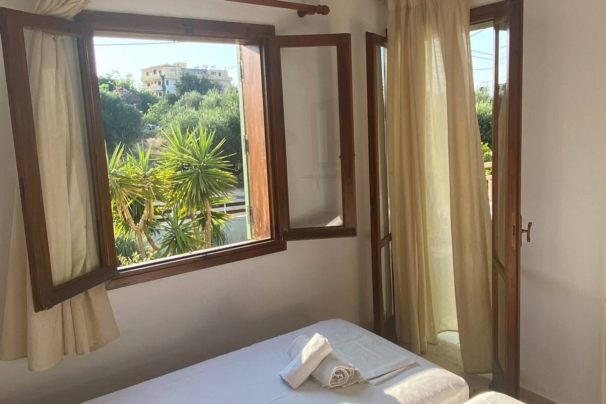 A bedroom with two beds, a window with curtains, a mirror, and a view of greenery outside.