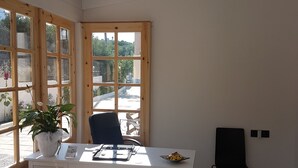 A room with a white desk, two teal ottomans, a black office chair, a teal pendant light, and a window with a view of the outside. - Sirocco Apartments (Hersonissos)