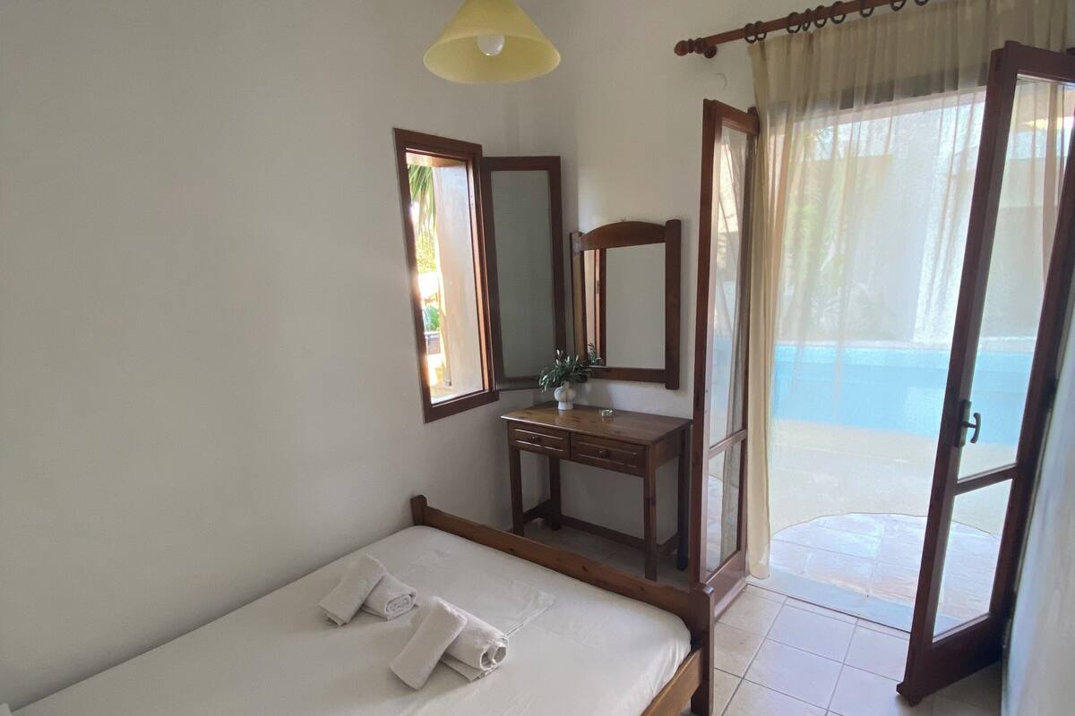 A bedroom with a bed, a desk, a mirror, and a view of the outside.