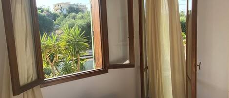 A bedroom with two beds, a window with curtains, a mirror, and a view of greenery outside.