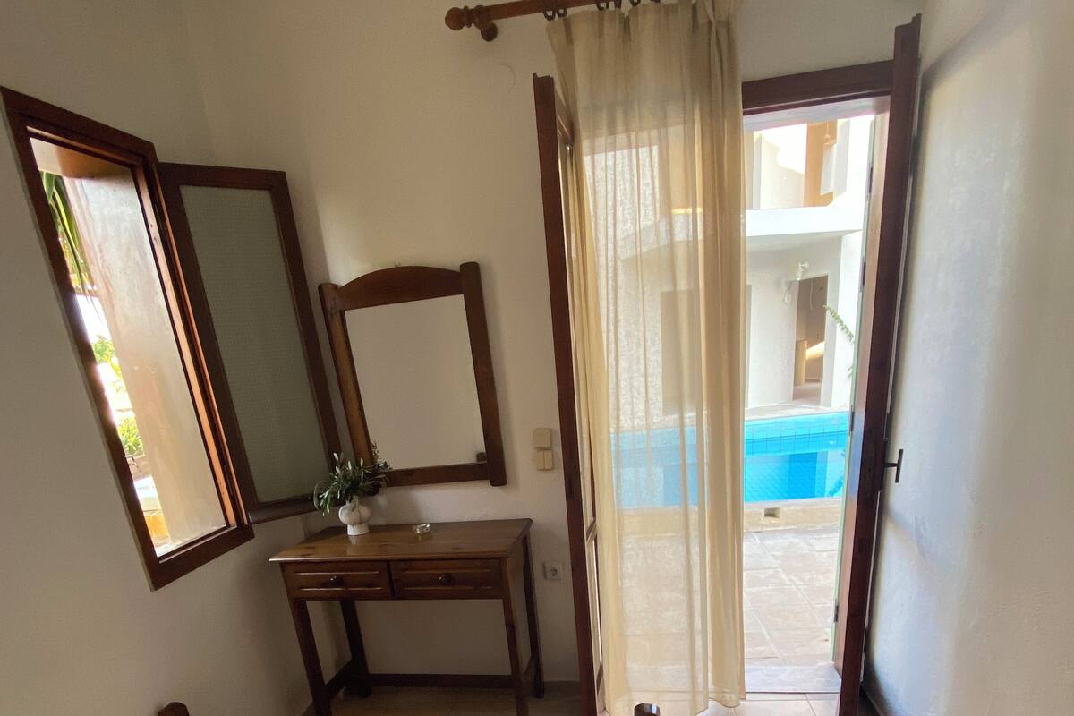 A bedroom with a bed, a desk, a mirror, and a view of a pool through a door.